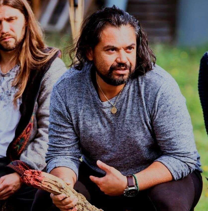 Man with long hair sitting outdoors, holding a wooden staff, wearing a gray sweater and necklace.